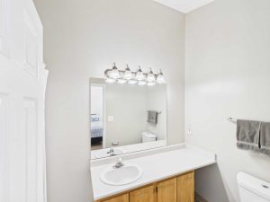 Apartments for rent in Limerick PA USA Bathroom with a single sink vanity, wooden cabinet, large mirror, five-bulb light fixture, toilet, and gray towel on a wall rack. Bedroom visible through open doorway.