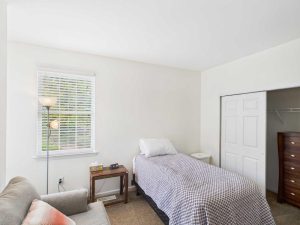 Apartments for rent in Limerick PA USA A simple bedroom with a single bed, side table, lamp, window with blinds, open closet with drawers, and part of a couch visible in the foreground.