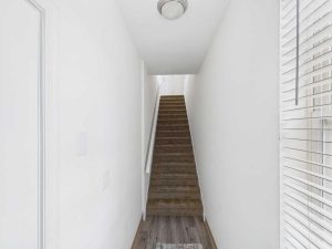 Apartments for rent in Limerick PA USA A narrow hallway with white walls and brown carpeted stairs leading up to a closed white door, with a window and blinds on the right.